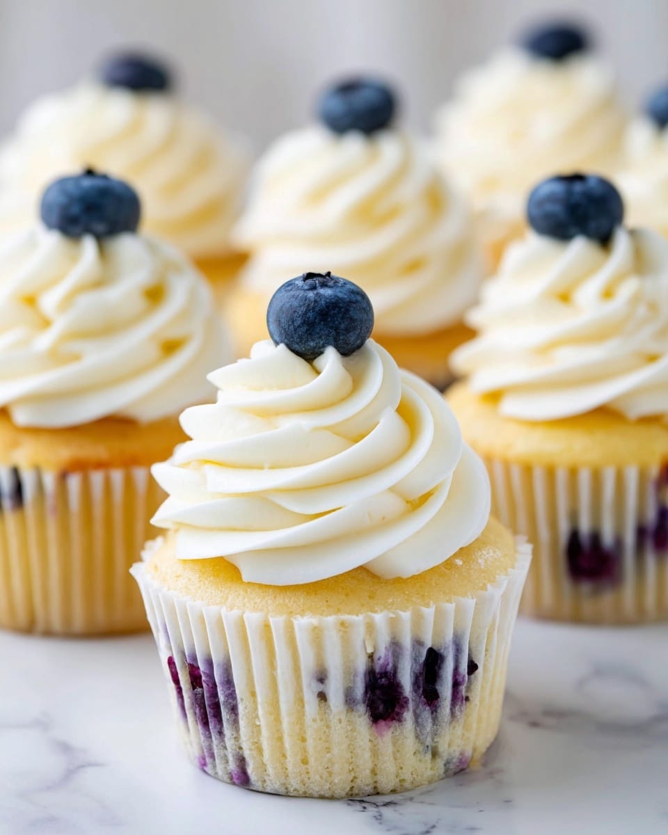 A close-up image of a blueberry cupcake with three distinct layers: the bottom layer is a pale yellow cake base with visible bits of deep purple blueberries baked inside, wrapped in a white paper liner with ridged edges. On top of the cake is a thick, soft layer of white creamy frosting swirled in smooth, rounded peaks, creating a textured, airy look. Two fresh dark blue blueberries sit on the side of the frosting, adding a pop of color and freshness. The cupcake is resting on a white plate with a blurred cluster of similar cupcakes in the background, all placed on a white marbled surface. photo taken with an iphone --ar 4:5 --v 7