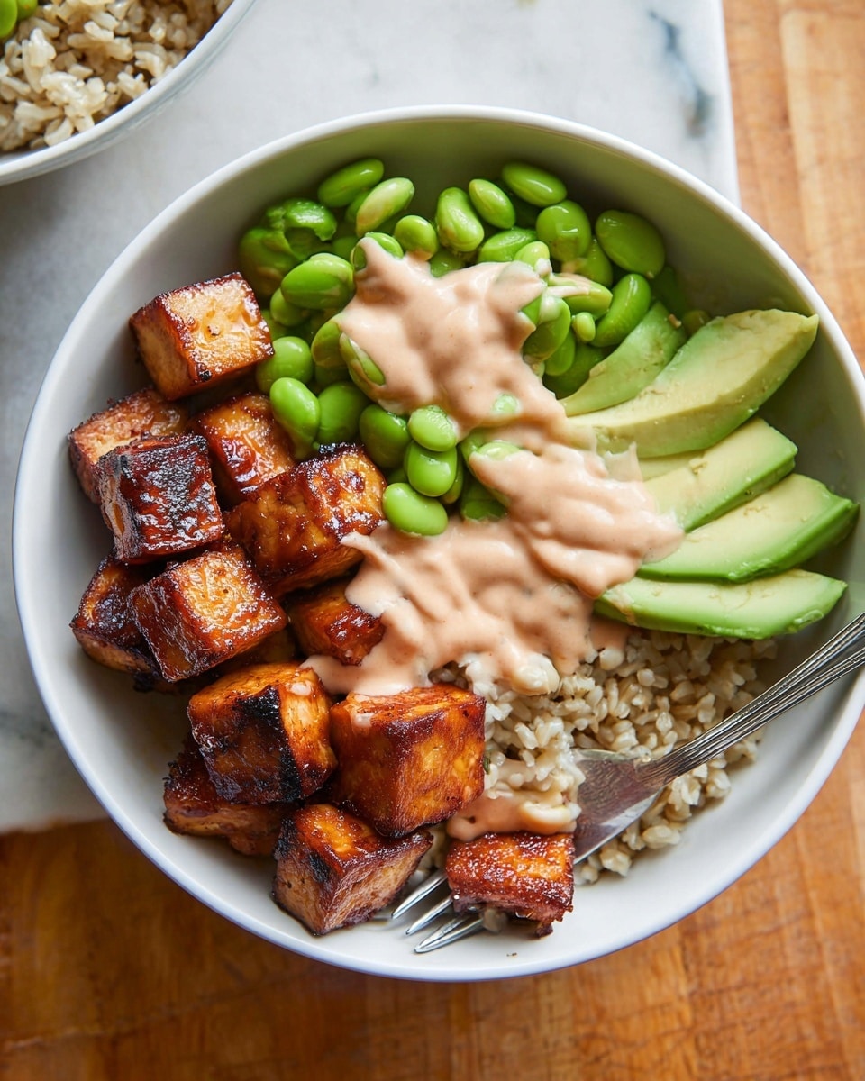 In a white bowl on a white marbled surface, the dish is arranged in four main parts: bright green edamame beans fill the upper left side, smooth light green slices of avocado fan out on the lower left side, and in the center beneath the other items is a bed of light brown rice. On the right side, there are dark brown glazed chunks of grilled tofu or meat stacked neatly. A creamy light pink sauce is drizzled over the tofu or meat pieces, avocado slices, and some of the edamame, adding contrast and texture. A silver fork rests inside the bowl at the bottom center. photo taken with an iphone --ar 4:5 --v 7