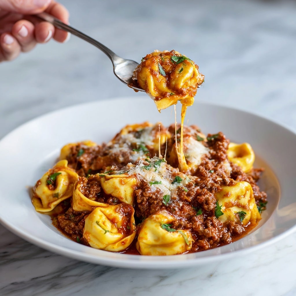 A white round plate holds a generous serving of tortellini pasta covered in a thick layer of rich, chunky meat sauce with visible bits of ground beef. The pasta is golden yellow with a slightly soft texture, tightly folded into small ring shapes. Melted white and light golden cheese is layered on top, slightly browned in spots, adding a creamy texture. Small green parsley sprigs are scattered over the dish, providing a fresh pop of color. To the right side of the plate rests a shiny silver fork. The plate is placed on a white marbled surface. photo taken with an iphone --ar 4:5 --v 7