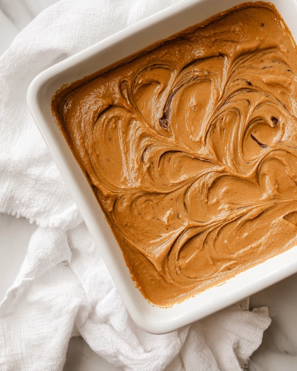 The image shows six square chocolate brownies arranged closely on white parchment paper over a white marbled surface. Each brownie has two visible layers: the bottom layer is thick, dark brown, and dense, while the top layer is a smooth, thick spread of light brown peanut butter frosting with a slightly glossy texture and swirling patterns. One brownie at the bottom right has a bite taken out, revealing the moist inside of the chocolate base with a few crumbs around it, and a small part of a knife is visible near it. photo taken with an iphone --ar 4:5 --v 7
