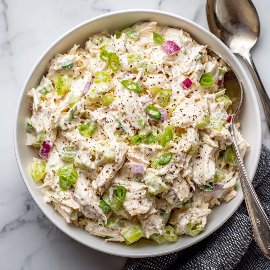 The image shows a close-up of a white bowl filled with a creamy chicken salad. The salad has shredded white chicken pieces mixed with small chunks of green celery, bits of red onion, and finely sliced green onions. The creamy dressing coats all the ingredients, giving the salad a smooth, slightly chunky texture with some specks of black pepper sprinkled on top. The bowl is placed on a white marbled surface with a small part of a silver spoon and a black-and-white cloth visible around it. photo taken with an iphone --ar 4:5 --v 7