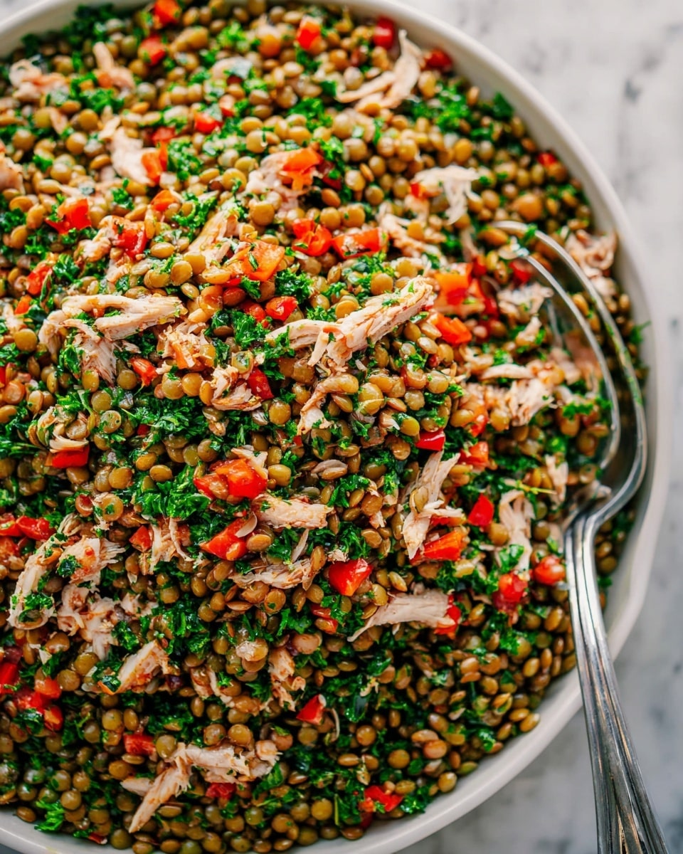 A close-up view of a lentil salad featuring three main layers: the base layer consists of small, round green lentils with a smooth texture, scattered evenly throughout the dish; the second layer includes finely chopped fresh green parsley, adding a leafy, textured look mixed all over; the top layer has chunks of light brown shredded chicken and diced bright red bell peppers, giving contrast in both color and shape. The dish is served in a white bowl with a silver spoon resting on the right side, all placed on a white marbled surface. Photo taken with an iphone --ar 4:5 --v 7